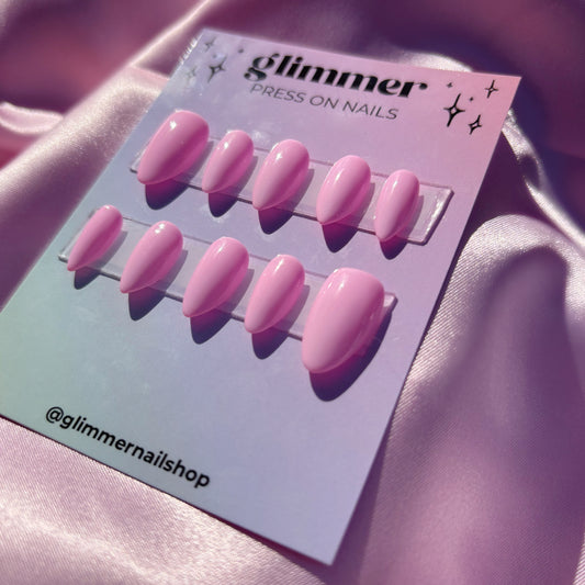 Pink press-on nails on a card with 'glimmer' branding against a pink fabric background