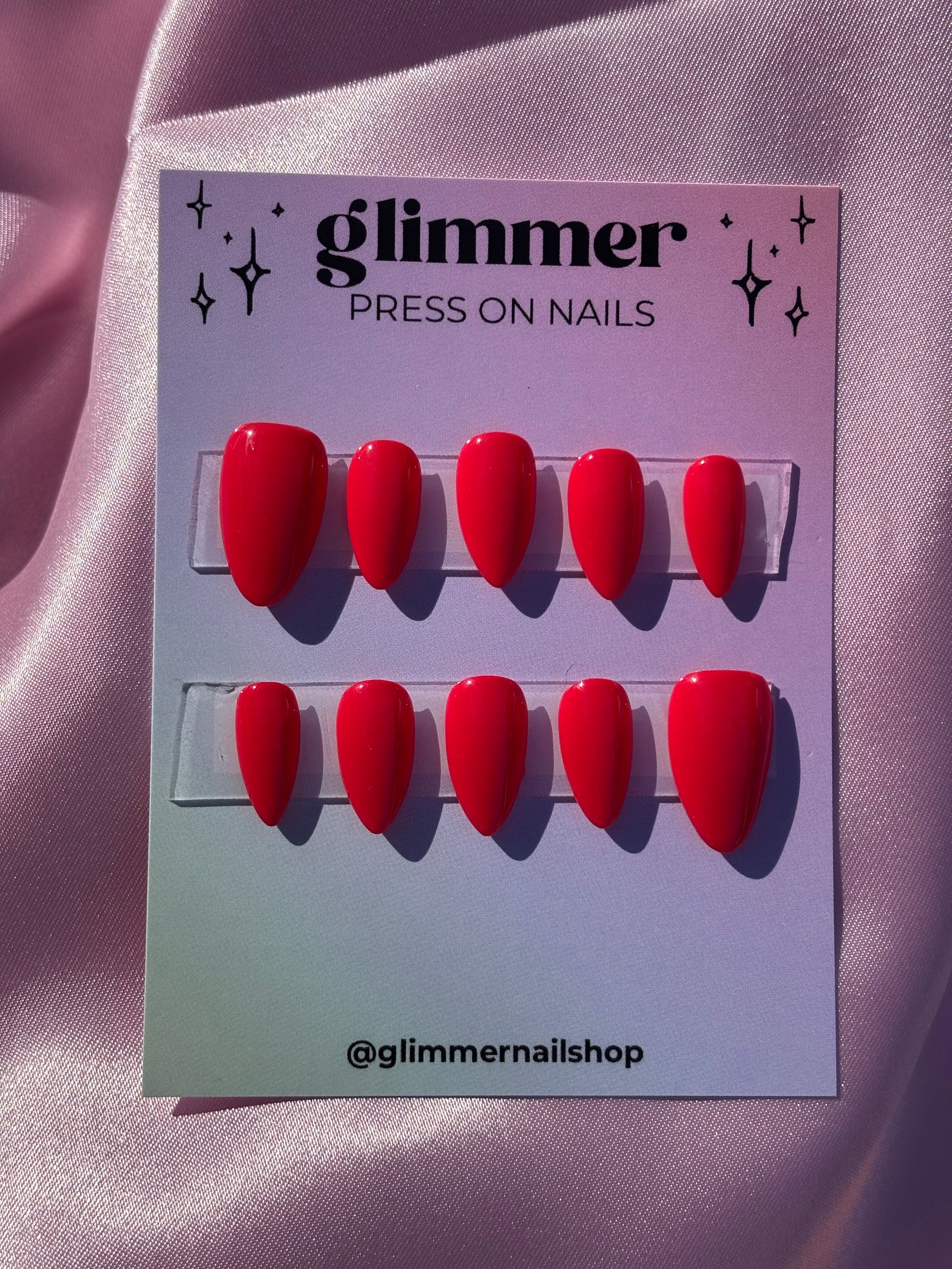 Red press-on nails on a card with 'Glimmer' branding on a pink satin background