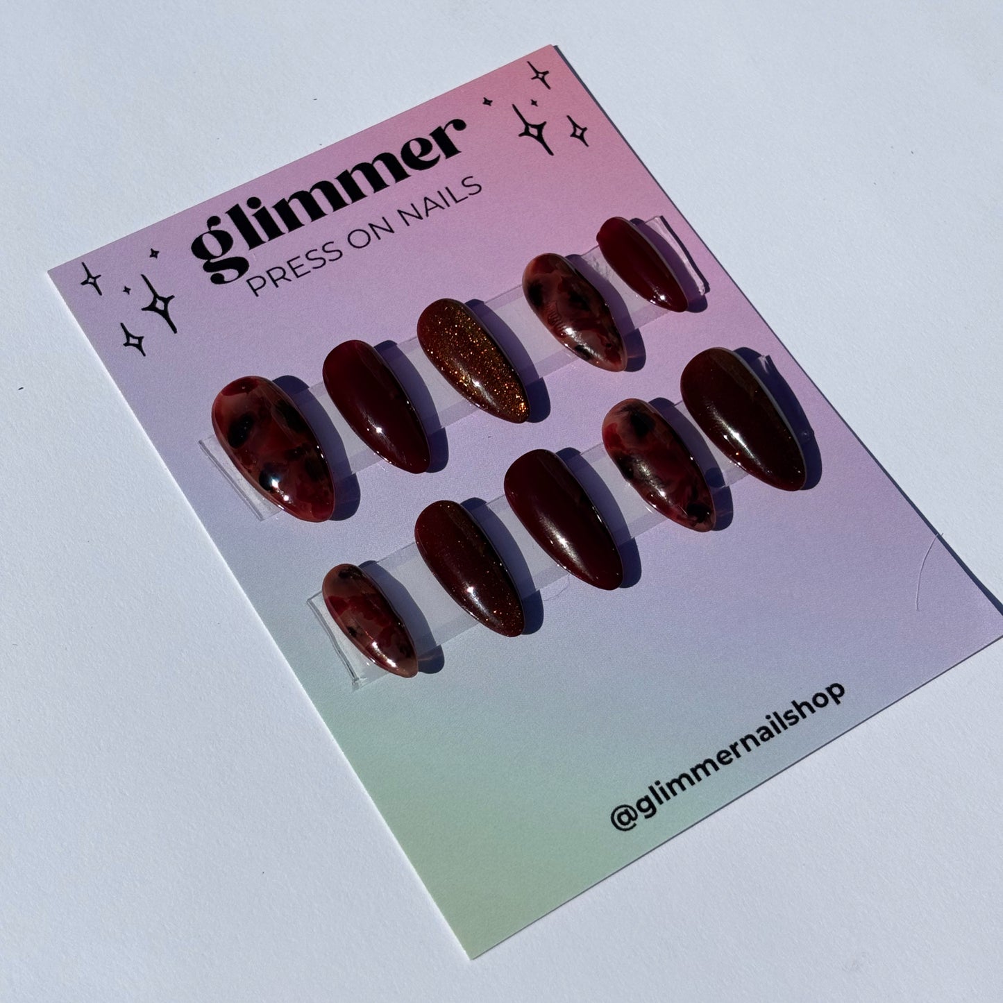 Set of burgundy press-on nails with glossy and glittery finishes, featuring various shapes and lengths.