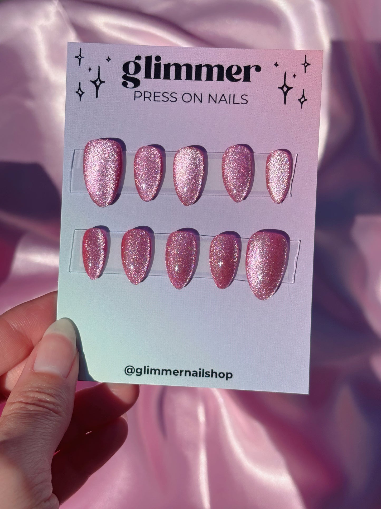 Card of glittery press-on nails held by a hand with a pink satin background