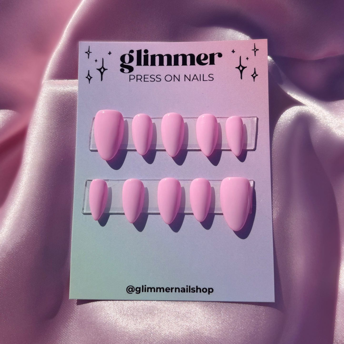 Pink press-on nails on a 'glimmer' branded card against a pink fabric background