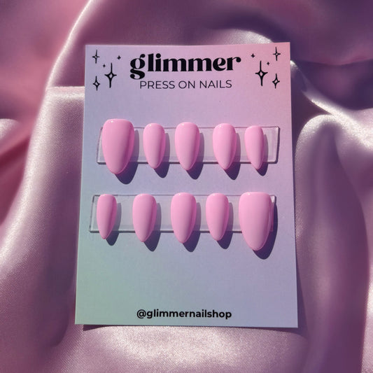 Pink press-on nails on a 'glimmer' branded card against a pink fabric background